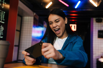 Photo of excited asian woman playing video game on mobile phone in cafe