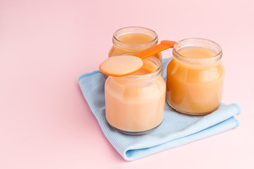 Glass jars of baby food with spoon and napkin on pink background
