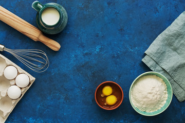 Obraz na płótnie Canvas Baking Ingredient- flour, egg, milk, rolling pin, whisk on a blue rustic background. Top view, flat lay, copy space.