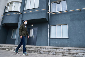 teen boy walks near wall of high-rise buildings with apartments, a residential area, a medical mask on his face protects against viruses and dust