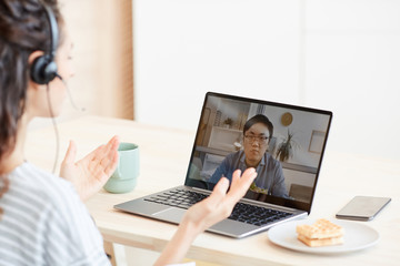 Young woman sitting at table in kitchen discussing something with her colleague using video call