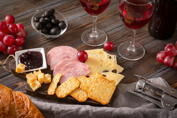 snack , 2 red wine glasses, slices of cheese , cracker, ham, olives, jam, grapes,  corkscrewon a dark wooden