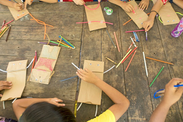 Top view of Asian kid & children group learning how to make paper puppet toy on wooden table background during free time or weaken, preschool or kindergarten daycare education concept, copy space 