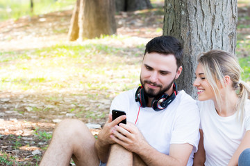 Happy couple young caucasian man using smartphone and headphones with woman leaning trunk of a tree together in the public park. Beautiful man and woman relaxing outdoors.Enjoying them favorite music.