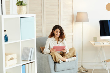 Young woman sitting relaxed in armchair and doing her job using modern tablet computer and Internet