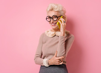 Grandma speaks on the phone. Photo of kind elderly woman wears eyeglasses on pink background.