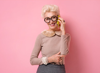 Grandma speaks on the phone. Photo of kind elderly woman wears eyeglasses on pink background.