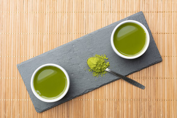2 bowls of organic matcha green tea in a bamboo napkin and stone slate. In a spoon, the green powder is slightly scattered. Top view