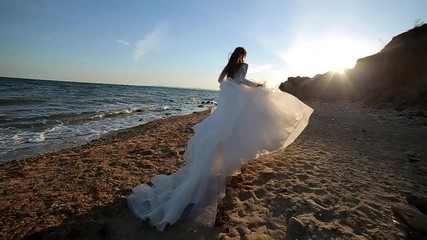 Attractive girl with long hair alone in a white wedding dress walks along the seashore. The bride runs at sunset. on blue sea background. Slow motion of woman, sexy woman, luxurious, smiling happy on 