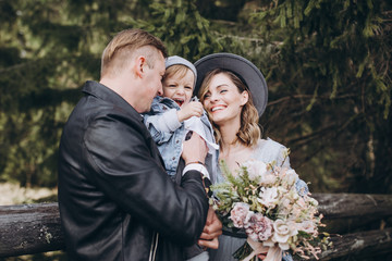 Stylish family in the mountains. A guy in a leather jacket, a girl in a wedding dress and a hat with his son stand under a fir tree near a wooden fence and hold a wedding bouquet