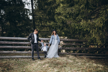 Fototapeta premium Stylish family in the mountains. A guy in a leather jacket, a girl in a wedding dress and a hat with his son stand under a fir tree near a wooden fence and hold a wedding bouquet