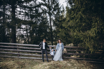 Stylish family in the mountains. A guy in a leather jacket, a girl in a wedding dress and a hat with his son stand under a fir tree near a wooden fence and hold a wedding bouquet