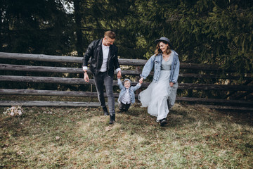 Fototapeta premium Stylish family in the mountains. A guy in a leather jacket, a girl in a wedding dress and hat run and play with his son against a background of green firs and a wooden fence