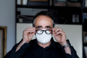 Ritratto di Uomo con occhiali da vista inossa una mascherina protettiva bianca , sullo sfondo l'interno di un appartamento, 