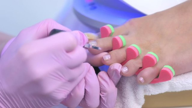 Pedicurist master is covering painting client's toes nails shellac, closeup. Another foot drying in UV led lamp. Professional pedicure in beauty salon. Hygiene care for hands. Beauty industry concept.