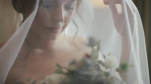 Close-up view of a charming pretty caucasian bride in a luxurious white wedding dress and veil holding a bridal bouquet of flowers behind a veil in the cozy bedroom of a hotel room in the morning.