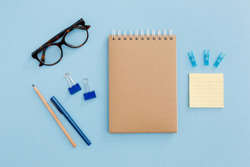 Blank brown vintage notebook, pencil, pen, diary, paper clips, glasses on colorful blue table. Stylish minimalistic workplace concept. Top view with copy space. 