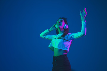 Listening to music. Caucasian young woman's portrait isolated on blue studio background in neon light. Beautiful female model. Concept of human emotions, facial expression, sales, ad, youth culture.