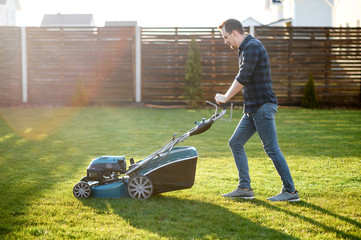Spring mowing the lawn. Young man in a plaid shirt and jeans mows grass with a lawn mower in the...