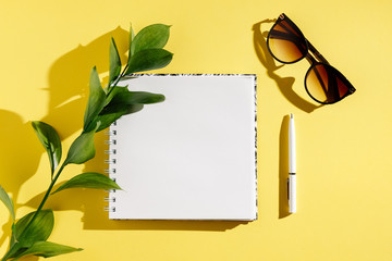 Blank white vintage notebook, leaves, diary, sunglasses on yellow  background. Minimalistic workplace concept. Top view with copy space. Summer flat lay