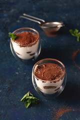 Tiramisu in a transparent glass with a double bottom with cocoa and mint on a blue dark background.
