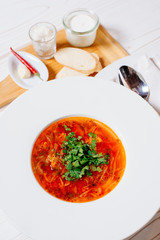 Borsch in a white plate on a white background, top view. Near puddles red pepper, garlic, sour cream and a spoon. Borsch - traditional Russian beet and cabbage soup.