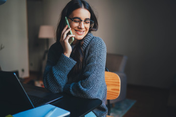 Happy young business woman in eyeglasses talking on cellphone laughing with colleague while working at home, Positive woman freelancer working remotely from home talking with friends on mobile phone