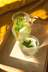 A small jug with lemon water and mint with a glass on a wooden tray in the interior with a yellow plaid. The concept of self-care and health.