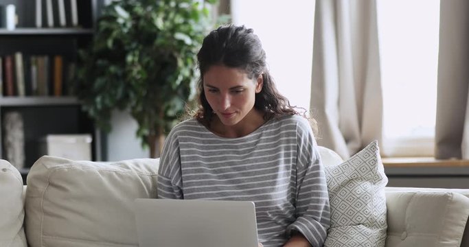Excited young lady winning online bid on laptop sitting on sofa at home. Overjoyed millennial woman winner looking at computer receiving email getting new remote job offer reading good news concept.