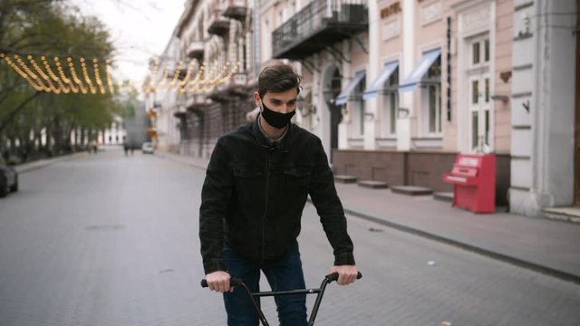 Portrait of young guy in protective face mask riding bike in the street of empty city center, slow motion