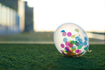A transparent balloon with a lot of colorful confetti on the inside laying on the grass with the sun and a blurry background horizontal