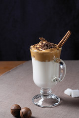 Glass mug of Dalgona coffee with chocolate crumb,  cinnamon stick, macadamia nuts and white lump sigar on brown tissue