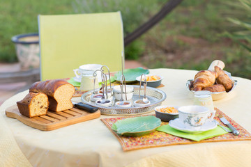 Homemade french brioche breakfast on a cutting board with homemade jam in cups and croissants. Green details, plates on the table outside on a sunny day in summer. Relaxing