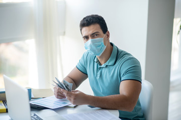 Dark-haired male sitting at his desk in protective mask, counting money