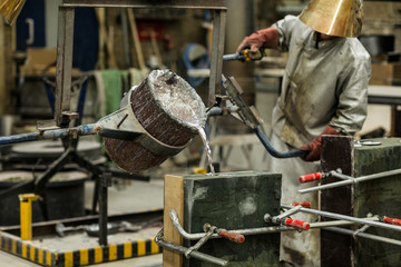 A craftsmen in silver protective clothing performing a metal sand casting technique by pouring...