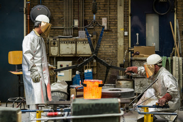 Two craftsmen in silver protective clothing doing metal sand casting technique pouring molten aluminum silver colored liquid into a mold in a industrial environment, workshop space