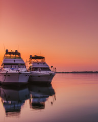 Two luxury yachts float moored against a dock with the calm lake waters and the pink, orange and...