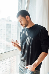 bearded man standing with hand in pocket and using smartphone near window at home
