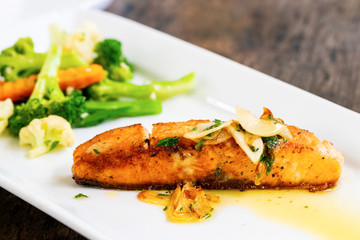 Close-up of garlic butter sauteed salmon on a white plate on a wooden table, soft focus on broccoli, cauliflower and carrots in the background
