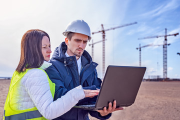 A builder and a real estate agent look into a laptop against the background of a building under construction.