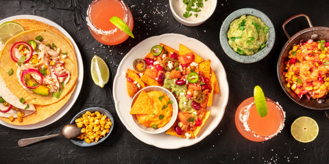 Mexican food, a flat lay panorama on a dark background. Nachos, tortillas, Paloma cocktails, guacamole and other dishes, shot from above