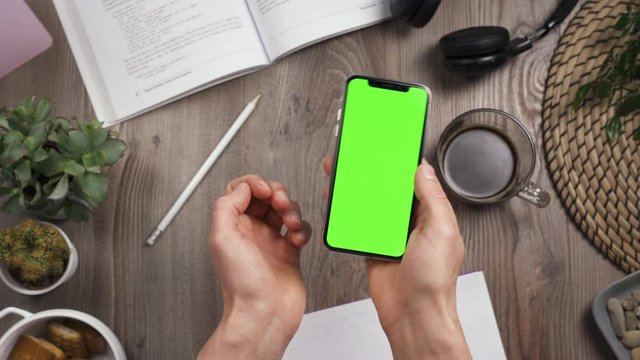 Male hands shake smartphone with green screen on it. Chroma key. Top view. Wooden table with many things on the background. Workplace table. Daylight.
