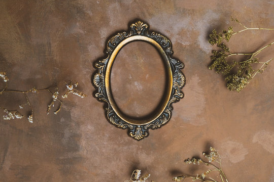 Empty oval bronze photo frame and twigs of dried flowers on decorative brown backdrop.