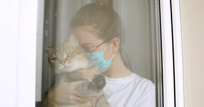 Girl in mask and glasses with cat is missing about usual normal life on quarantine at home looking at window. Depression in self-isolationg during covid-10 pandemic, epidemic. Portrait of woman.