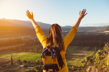 Young woman stands at the top of a hill with her arms raised at golden hour