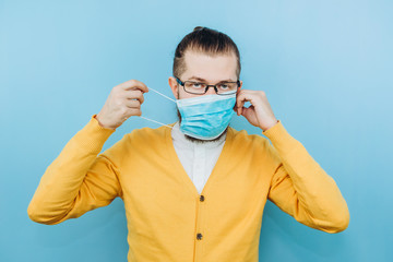 A young man in a medical mask on a blue background. A man in a yellow cardigan shows a hand gesture OK. We're going to beat coronavirus. The COVID-2019 epidemic. Positive boy.