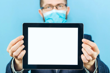 A man in a suit, glasses and a medical mask holds a piece of paper, on a blue background. Place for text. Businessman holds a tablet in his hands. The problem of business due to coronavirus. COVID-19