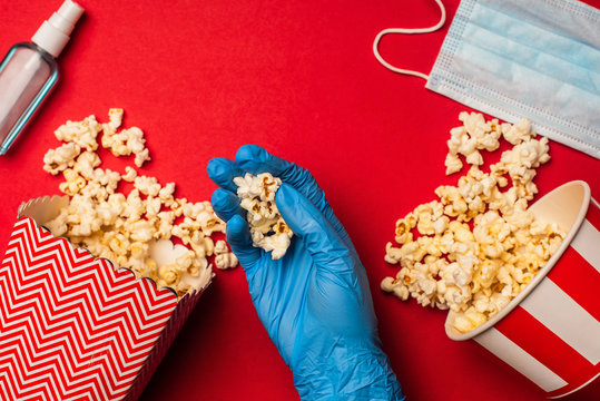 Top view of person in latex glove holding popcorn near hand sanitizer and medical mask on red background