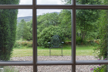 through a grid created by a wooden window we can see an ornate iron chair in the garden of the house