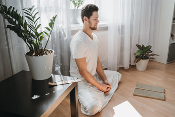 Young man meditating on his living room on the floor, sitting in the lotus position with closed eyes. Expression of tranquility in a health, mindfulness and fitness concept.